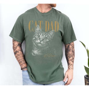 May include: An olive green short-sleeve t-shirt featuring the text CAT DAD in gold lettering above a detailed black and white sketch of a tabby cat. Below the cat illustration, the word Katrina is printed in a cursive gold script font.