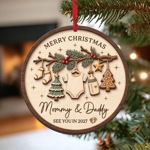 Puede incluir: Un adorno navideño redondo de madera con borde oscuro y centro claro. Muestra una rama con artículos de bebé colgando, incluyendo un body, biberón, calcetines y árboles. El texto dice MERRY CHRISTMAS, Mommy & Daddy, SEE YOU IN 2027.