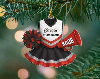 May include: A cheerleader uniform ornament featuring a white top with red and silver stripes, a black skirt with red and silver trim, a black and red pom-pom, and a red megaphone with the text 2026. The top displays the text Caryn and Team Name.