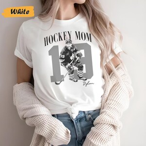 Może przedstawiać: Biała koszulka z napisem "HOCKEY MOM" nad szarym zdjęciem hokeisty i numerem 19. Numer 19 jest duży i szary, ze zdjęciem gracza pośrodku. Poniżej widnieje imię "Dylan".