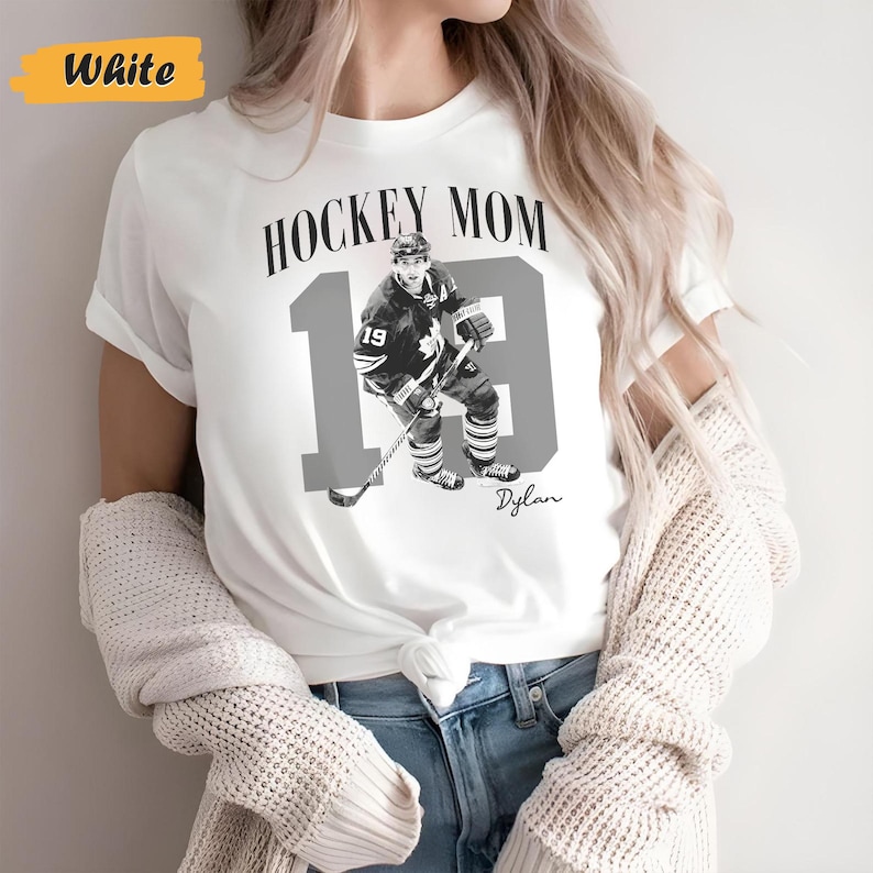 Może przedstawiać: Biała koszulka z napisem "HOCKEY MOM" nad szarym zdjęciem hokeisty i numerem 19. Numer 19 jest duży i szary, ze zdjęciem gracza pośrodku. Poniżej widnieje imię "Dylan".