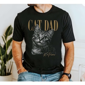 May include: A black short-sleeved t-shirt featuring a graphic of a tabby cat in the center. Above the cat, the text CAT DAD is printed in a gold-toned font. Below the cat, the name Katrina is printed in a gold-toned cursive script font.
