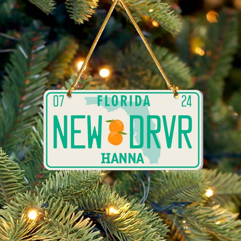 Puede incluir: Un adorno de placa de matr&iacute;cula de Florida blanco y verde con el texto "NEW DRVR" y el nombre "HANNA". El adorno presenta un peque&ntilde;o gr&aacute;fico naranja y el contorno del estado de Florida.