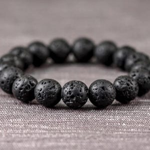 Puede incluir: Una pulsera circular compuesta por numerosas cuentas esféricas de piedra volcánica negra. Cada cuenta presenta una superficie porosa y texturizada con pequeñas hendiduras y un acabado mate, ensartadas para formar un bucle continuo.