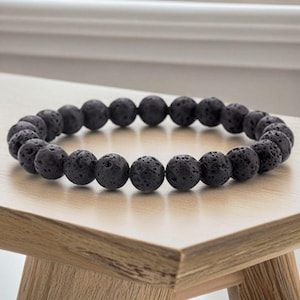 Puede incluir: Una pulsera elástica circular compuesta por numerosas cuentas esféricas de lava negra. Cada cuenta presenta una superficie porosa y texturizada característica de la roca volcánica, dispuesta en un bucle continuo para formar un accesorio.