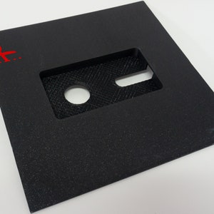 Puede incluir: Una pieza cuadrada de material negro con un recorte en forma de tarjeta de crédito. El recorte tiene un pequeño círculo en el centro.