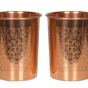 Puede incluir: Dos vasos de color cobre con un diseño floral grabado. Los vasos tienen forma cilíndrica con un borde ligeramente ensanchado. El patrón grabado cubre la mayor parte de la superficie, añadiendo un elemento decorativo.