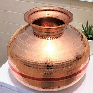 Puede incluir: Un jarrón redondo de color cobre con una abertura ancha y una banda decorativa. El jarrón tiene una textura martillada y una banda marrón rojiza en el medio. El jarrón está sobre una superficie blanca.