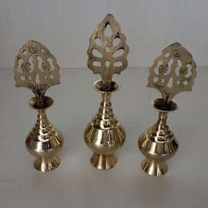 Puede incluir: Tres botellas decorativas de latón con tapas caladas y ornamentadas. Las botellas tienen forma bulbosa con base acampanada y una serie de estrías. Las tapas tienen forma de hojas o diseños florales estilizados.