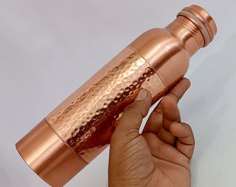May include: A copper water bottle with a hammered texture on the body. The bottle has a cylindrical shape with a screw-on cap. The copper has a shiny, reddish-brown color. The bottle is held by a hand.
