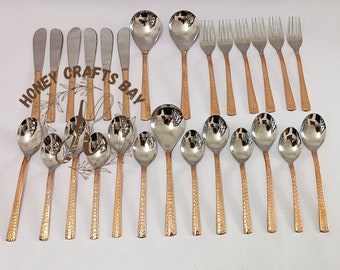 May include: A collection of stainless steel and copper-toned flatware, including spoons, forks, and butter knives. The spoons have large, rounded bowls, and the handles feature a hammered texture. The flatware is arranged on a white surface.
