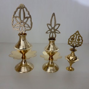 May include: Three decorative items in a gold colour. They have a tiered, vase-like form with detailed floral or leaf-like designs at the top. The bases have a scalloped edge. The items vary in size, with the tallest one being approximately 15 cm tall.