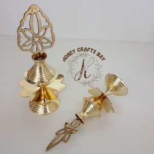 Pode incluir: Duas lamparinas de óleo de metal dourado com desenhos intrincados. As lamparinas têm uma base redonda e um pescoço alto e fino com uma parte superior decorativa. A parte superior da lamparina tem a forma de uma flor com um furo no centro. As lamparinas são feitas de latão e têm um acabamento brilhante.