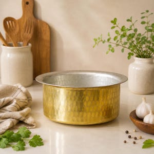 Puede incluir: Una olla de cocina de latón martillado con borde plateado. La olla está sobre una superficie clara, con una tabla de cortar de madera, utensilios y un pequeño jarrón con vegetación en el fondo. Ajo y granos de pimienta en un cuenco de madera.
