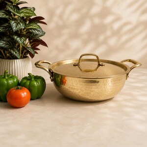 Puede incluir: Una olla redonda de latón martillado con dos asas laterales y una tapa a juego con un asa central. La olla tiene un acabado dorado pulido y reflectante con una superficie texturizada. Está colocada junto a dos pimientos verdes y un tomate rojo.