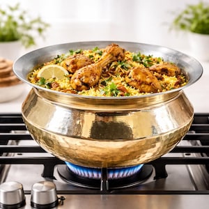 May include: A traditional brass cooking pot filled with chicken biryani, garnished with fresh herbs and a lemon slice. The pot is on a gas stove with a blue flame. The pot has a silver rim. The dish is being cooked on a gas hob.
