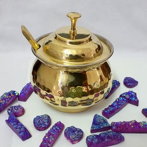 Puede incluir: Un azucarero de metal dorado martillado, con tapa y cuchara. El cuenco está rodeado de varias formas de formaciones de cristal morado y azul, creando una exhibición decorativa y elegante.