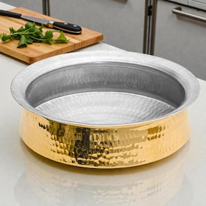 May include: A hammered metal serving bowl with a gold-colored base and a silver-colored rim. The bowl has a wide, shallow design and a textured surface. A wooden cutting board with a knife and herbs is in the background.