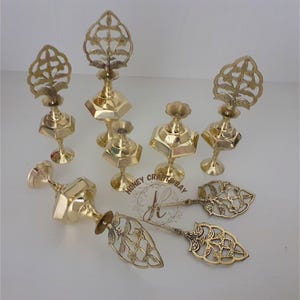 May include: A collection of ornate, gold-colored metal decorative items. The assortment includes several candle holders with intricate floral or leaf-like designs, and two serving spoons with similar decorative patterns. The items are arranged on a light-colored surface.