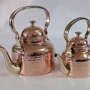 Puede incluir: Dos teteras de cobre con asas y boquillas de latón. Las teteras tienen una textura martillada y un acabado brillante. La más grande mide unos 25 cm de alto, y la más pequeña unos 18 cm. Ambas tienen un diseño clásico.