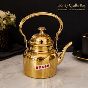 May include: A polished gold-colored brass kettle featuring a rounded body, a curved spout, and a high arched handle. A small white rectangular sticker with the red text BRASS is affixed to the front of the kettle, which has a smooth, reflective surface.