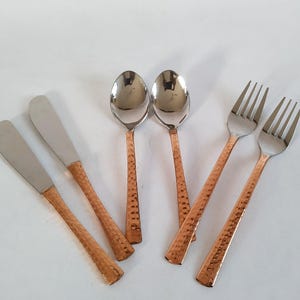 Könnte beinhalten: Ein Set aus Edelstahlbesteck mit kupferfarbenen, gehämmerten Griffen. Das Set umfasst zwei Buttermesser, zwei Löffel und zwei Gabeln. Die Löffel und Gabeln haben eine glänzende Oberfläche, während die Messer eine matte Oberfläche haben.