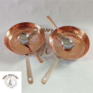 May include: Two copper-colored pans with hammered textures and silver-colored handles. Each pan has a matching silver-colored spoon resting inside. The pans are round with a small spout on one side. The image is of kitchenware.