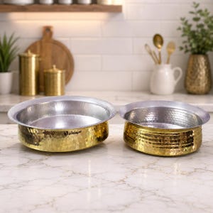 Könnte beinhalten: Zwei gehämmerten Metallschalen mit goldener Außenseite und silberner Innenseite stehen auf einer Marmoroberfläche. Die Schalen haben ein breites, flaches Design, ideal zum Servieren von Speisen. Die strukturierte Goldoberfläche verleiht dem Geschirr Eleganz.