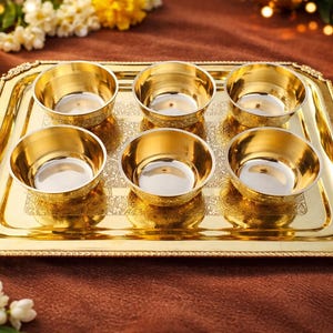 May include: A gold-coloured serving tray with six small, round bowls. The tray and bowls have a shiny finish. The tray has a decorative border and is set on a brown surface. Flowers and a lit candle are in the background.