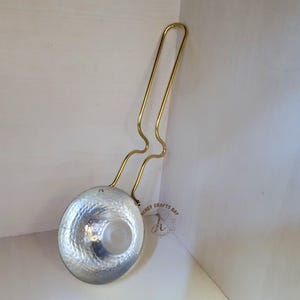 Puede incluir: Un cucharón plateado con un cuenco martillado y un mango dorado largo y curvado. El mango está unido al cuenco con un diseño decorativo. El cucharón está apoyado contra una superficie blanca.