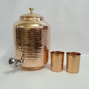 以下が含まれることがあります： ハンマー加工された銅製のウォーターディスペンサー。蛇口、蓋、お揃いの銅製カップ2個付き。ディスペンサーは丸い形で、表面はテクスチャード加工されています。カップは円筒形で、ハンマー加工されたテクスチャも施されています。