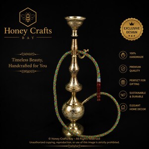 May include: A gold-colored metal hookah featuring intricate engraved patterns on the base and stem. It includes a long, flexible hose with a multi-colored woven pattern and a dark brown handle. The hookah is displayed against a black background.