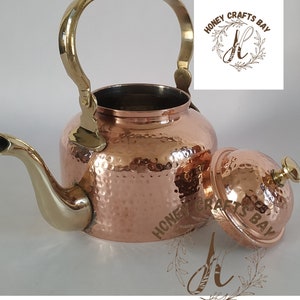 May include: A copper tea kettle with a hammered texture, featuring a brass handle, spout, and lid. The lid is slightly ajar, revealing the interior. The kettle has a classic design, suitable for kitchen decor.