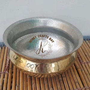 May include: A silver and gold hammered metal bowl with the text "Honey Crafts Bay" and a stylised letter "H" inside a wreath. The bowl is sitting on a woven bamboo mat.