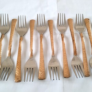 Puede incluir: Diez tenedores con un diseño de dos tonos. Las púas y el cuenco son plateados, mientras que los mangos tienen un acabado de cobre martillado. Los tenedores están dispuestos sobre una superficie blanca.