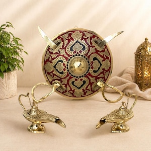 May include: A decorative set featuring a red and gold shield with two swords, and two gold genie lamps. The shield has an ornate design. The lamps have curved spouts and handles. A gold lantern is in the background.