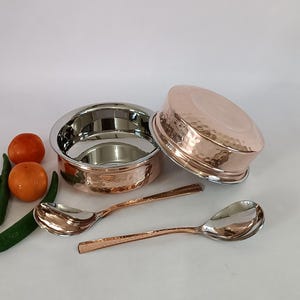 Puede incluir: Cuencos y cucharas de servir de color cobre. Los cuencos tienen una textura martillada en el exterior y un interior plateado brillante. Se ven dos cucharas, una con mango de cobre martillado y una cabeza de cuchara plateada.