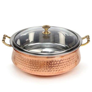 May include: A hammered copper serving dish with a glass lid and brass handles. The dish has a hammered texture and a round shape.
