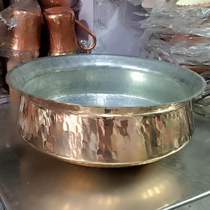 Puede incluir: Un cuenco grande de cobre martillado con un diseño ancho y poco profundo. El cuenco tiene una superficie brillante y reflectante y una textura martillada en el exterior. El interior es de un color plateado más claro. Otros utensilios de cocina de cobre son visibles en el fondo.