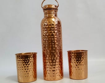 Könnte beinhalten: Ein Set aus drei Kupfer-Trinkgefäßen mit gehämmerter Textur. Das Set enthält eine hohe Flasche mit Deckel und zwei kürzere Becher.