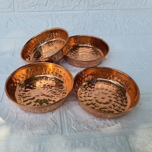 May include: Four copper-coloured bowls with a hammered texture. The bowls are round with a slightly flared rim. The interior of the bowls has a hammered design. The bowls are arranged on a white surface.