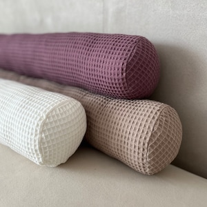 Könnte beinhalten: Drei zylindrische Kissen mit Waffelstruktur in verschiedenen Farben: Lila, Weiß und Beige. Die Kissen sind übereinander gestapelt.