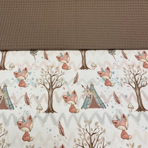 Könnte beinhalten: Ein weißer Stoff mit einem sich wiederholenden Muster aus braunen Bäumen, rosa und blauen Zickzacklinien und Füchsen, die Kopfbedeckungen tragen. Die Füchse sitzen und haben braunes Fell, weiße Akzente und rosa Nasen. Der Stoff ist mit einem braunen Waffelstoff überzogen.