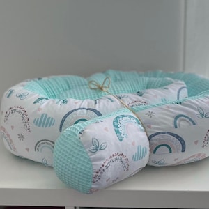 Könnte beinhalten: Ein türkisfarbenes und weißes Baby-Nest mit einem Regenbogenmuster. Das Nest ist aus weichem Stoff gefertigt und hat ein abnehmbares Kissen.