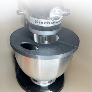 Könnte beinhalten: Ein silberner Küchenmixer mit schwarzem Sockel und einer schwarzen Schüssel mit grauem Deckel. Der Deckel hat eine kleine Öffnung zum Einfüllen von Zutaten. Der Mixer trägt den Schriftzug "KitchenAid" auf der Seite.