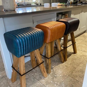 May include: Three leather bar stools with wooden legs and a black metal footrest. The stools are teal, brown, and black.