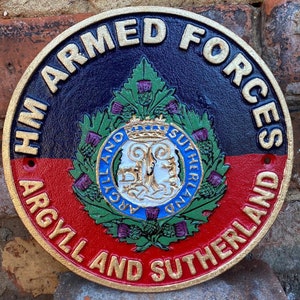 May include: A round, cast iron sign with gold lettering that reads "HM Armed Forces Argyll and Sutherland". The sign features a blue and red background with a crest in the centre. The crest is surrounded by green leaves and purple thistles.