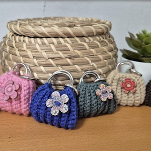 Puede incluir: Seis pequeños bolsos de mano de ganchillo en varios colores, incluyendo rojo, rosa, azul y marrón. Cada bolso tiene una flor decorativa y un asa plateada. Una cesta tejida y plantas en macetas se encuentran en el fondo.