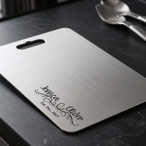 May include: A rectangular, brushed stainless steel cutting board with a handle cutout. The board features the script text "Jessica Oliver Est. Dec, 2025" in black. A fork and spoon rest on the dark surface.