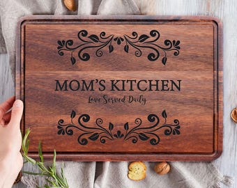 May include: A rectangular wooden cutting board with a dark brown finish. The text "MOM'S KITCHEN Love Served Daily" is engraved in the center, with decorative floral designs above and below. The board is held by a hand.